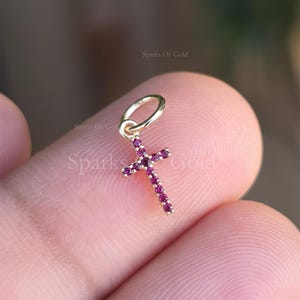 Op de afbeelding: Een kleine gouden kruis hanger met een lus voor een ketting. Het kruis is versierd met kleine, ronde, magenta gekleurde edelstenen. De hanger wordt tussen twee vingers gehouden, met de tekst "Sparks Of Gold" zichtbaar.