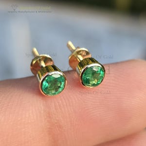 Könnte beinhalten: Ein Paar goldene Ohrstecker mit runden, leuchtend grünen Smaragden. Die Ohrringe haben ein schlichtes, elegantes Design, wobei die Smaragde in einer goldenen Lünette gefasst sind. Die Ohrstecker sind klein und zierlich.