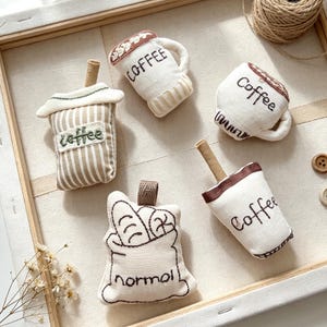Könnte beinhalten: Fünf Stoffornamente mit gestickten Motiven zum Thema Kaffee und eine Tüte mit Brot. Drei Artikel sind als Kaffeetassen mit dem gestickten Wort "Coffee" geformt, einer ist ein gestreifter Kaffeebecher und einer ist ein Beutel mit der Aufschrift "normal".