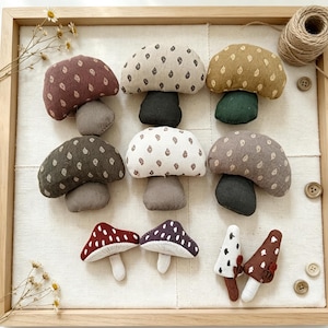 Può includere: Una collezione di undici funghi in tessuto imbottito di vari colori, tra cui bordeaux, crema, giallo senape, verde oliva, marrone e viola scuro, con motivi a piccole foglie o puntini, disposti su una superficie in tessuto chiaro in una cornice di legno.
