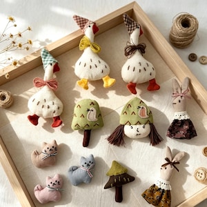 May include: A collection of small fabric ornaments including three white geese with patterned hats and bows, two rabbits in dresses, three cats, two trees, and a doll head with brown hair, all arranged on a light fabric surface inside a wooden tray.