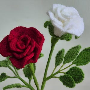 Może przedstawiać: Szydełkowe róże w kolorze czerwonym i białym z zielonymi łodygami i liśćmi. Ręcznie robione kwiaty mają szczegółowe płatki i realistyczne liście, idealne do dekoracji domu lub na prezent.