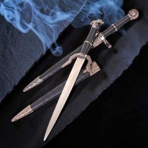 May include: A double-edged dagger with a silver-toned blade, a black grip, and ornate metallic accents on the hilt and pommel. The dagger is accompanied by a matching black scabbard featuring intricate silver-toned metal detailing at the tip and throat.