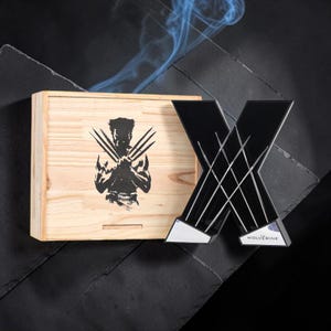 May include: A light-coloured wooden box featuring a black silhouette of a character with three extended claws, paired with a black X-shaped display piece. The display piece has silver claw marks across its surface and the text THE WOLVERINE.