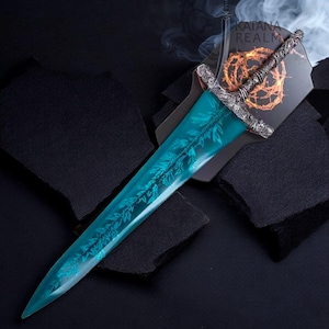 May include: A decorative sword with a teal blade featuring a leaf design. The hilt is silver with intricate detailing. The sword rests on a black surface, with the text "KATANA REALM" visible on the backing.