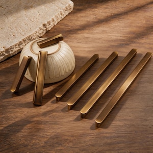 May include: A selection of antique brass cabinet pulls and a small decorative bowl. The pulls are rectangular with rounded ends. The bowl is off-white with a textured surface. The items are arranged on a wooden surface.