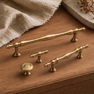 May include: A set of antique brass cabinet hardware, including two handles, two smaller pulls, and a knob. The hardware has a classic, ornate design with a warm, golden finish. The pieces are arranged on a wooden surface.