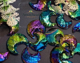 Op de afbeelding: Iridescent halvemaanvormige hangers in tinten paars, blauw, groen en goud. De hangers zijn verspreid over een donkergrijze ondergrond, met witte bloemaccenten op de achtergrond. Het logo "Arrowhead Hub" is zichtbaar.