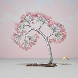 May include: A decorative tree sculpture with a silver-coloured metal trunk and branches. The branches are adorned with pink gemstones, resembling blossoms. The tree is mounted on a small, rectangular base. The background is a soft pink.