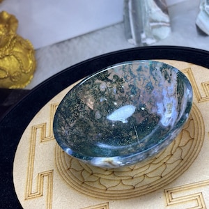 Könnte beinhalten: Eine polierte, ovale Schale aus Moosachat mit grünen und weißen Mustern. Die Schale steht auf einem hellen, lasergeschnittenen Holzsockel mit geometrischem Design. Im Hintergrund ist ein goldfarbenes Dekorationsobjekt zu sehen.