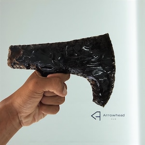May include: A dark obsidian axe head, held in a hand, against a white background. The axe head is roughly hewn with a polished, reflective surface. The logo for Arrowhead Hub is in the lower right corner.