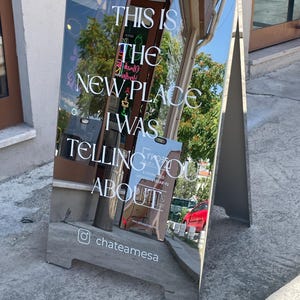 May include: A silver-toned, A-frame pavement sign with a reflective surface. The sign features white text that reads: THIS IS THE NEW PLACE I WAS TELLING YOU ABOUT, followed by an Instagram icon and the handle chateamesa at the bottom.