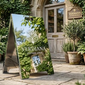 Puede incluir: Un letrero plateado en forma de A con el logotipo "Verdiana Boutique Jardin" en blanco. El letrero refleja la vegetación y el edificio circundantes. Está colocado sobre un patio de piedra frente a una puerta de madera y plantas en macetas.