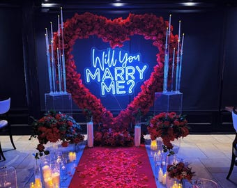 Op de afbeelding: Een romantische setting voor een aanzoek met een groot bloemstuk in de vorm van een hart. Een neonbord toont "Will You MARRY ME?" boven een rode loper bezaaid met rozenblaadjes. Hoge kaarsen en bloemboeketten maken de scène compleet.