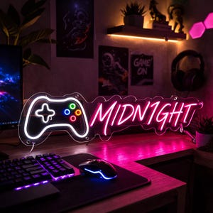 Op de afbeelding: Een neonbord in de vorm van een gamecontroller met het woord "MIDNIGHT" in roze neon. De controller heeft een witte omtrek en gekleurde knoppen. Het bord staat op een bureau met een toetsenbord en muis.