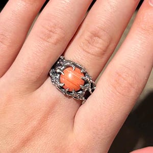Könnte beinhalten: Ein silberner Ring mit einem ovalen, korallenfarbenen Stein. Der Ring hat ein detailliertes, strukturiertes Design um den Stein herum. Der Ring befindet sich an einer Hand mit langen, hellrosa Fingernägeln.