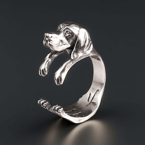 Könnte beinhalten: Ein silberner Ring mit einem detaillierten Hundedesign. Der Ring zeigt eine Skulptur des Kopfes und der Vorderpfoten eines Hundes. Der Ring bildet den Körper des Hundes. Ein einzigartiges Schmuckstück.