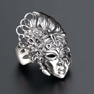 May include: A silver-toned ring featuring a detailed mask design. The mask has intricate facial features, eye cutouts, and ornate embellishments. The ring's design includes a decorative headpiece with floral and scrollwork elements.