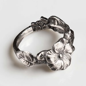 Könnte beinhalten: Ein silberfarbener Ring mit floralem Design. Der Ring hat ein Blumen- und Blättermotiv mit detaillierten Texturen. Der Ring ist vor einem weißen Hintergrund platziert, der die filigranen Details des Schmucks hervorhebt.