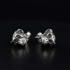 Puede incluir: Un par de pendientes de plata, cada uno con la forma de una cabeza de animal estilizada con ojos y hocico visibles. Los pendientes están sobre un fondo oscuro, resaltando su brillo metálico y detalles intrincados. Probablemente hechos de plata de ley.