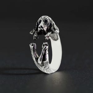 Könnte beinhalten: Ein silberner Ring mit detailliertem Hundekopf- und Pfoten-Design. Der Hundekopf befindet sich oben, die Pfoten an den Seiten. Der Ring hat eine polierte Oberfläche und ein einzigartiges Design. Ein charmantes Schmuckstück.