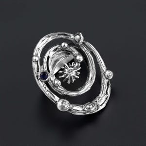 Könnte beinhalten: Ein silberfarbener Ring mit einem wirbelnden, organischen Design. Der Ring hat ein zentrales sonnenstrahlenartiges Element und einen kleinen, dunkelblauen Edelstein. Das Design enthält mehrere kugelförmige Akzente.