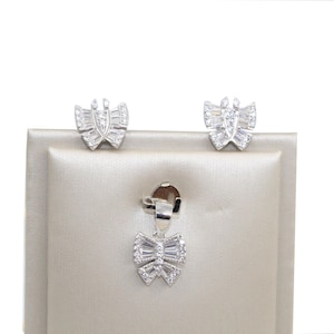 May include: A jewelry set consisting of two stud earrings and a pendant, each shaped like a bow. The pieces are crafted from a silver-toned metal and are encrusted with clear, baguette-cut and round-cut stones that create a sparkling effect.