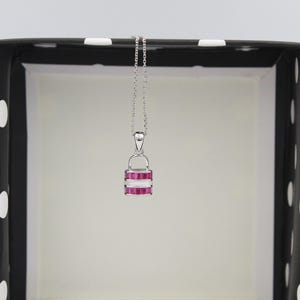 May include: A silver necklace with a pendant featuring a stacked design of pink gemstones and white accents. The pendant hangs from a delicate chain. The necklace is displayed against a black and white polka dot background.
