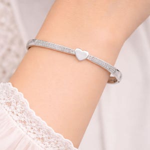 Pode incluir: Uma pulseira prateada com um berloque em forma de coração. A pulseira apresenta uma fileira de pequenas pedras brilhantes ao longo da banda. O berloque de coração está centrado na pulseira. A joia é usada no pulso.
