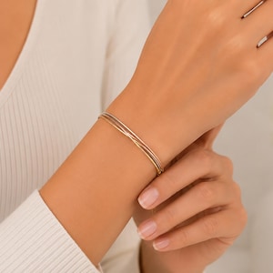 May include: A delicate tri-colour bangle bracelet featuring three thin, intertwined metal bands in yellow, rose, and white gold tones. The bracelet has a smooth, polished finish and a slender, circular design that rests comfortably on the wrist.
