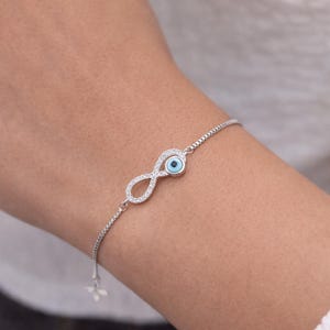 Peut inclure: Un bracelet en argent orné d'un symbole infini serti de cristaux transparents. Il comprend également un petit charme en forme d'œil bleu clair. La chaîne réglable est délicate et le bracelet est porté au poignet.