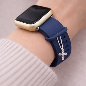 Może przedstawiać: Złoty smartwatch z granatowym silikonowym paskiem. Srebrny charms z krzyżem wisi na pasku. Zegarek ma czarny ekran.