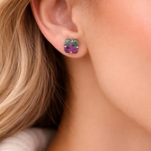 Puede incluir: Pendientes cuadrados con aspecto de piedra natural. Los pendientes presentan una combinación de tonos verdes y magenta, engastados en un metal color plata. Los pendientes se muestran en una oreja.