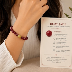 Peut inclure: Un bracelet composé de perles rondes polies rouge foncé et d'une perle en métal doré en forme de tête de Bouddha. À côté se trouve une carte rectangulaire avec le texte RUBY JADE STONE OF PASSION & PROTECTION et une liste de bienfaits.