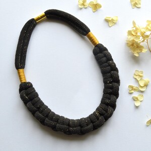 Peut inclure: Un collier noir tressé avec des détails dorés. Le collier est fait d'un matériau épais et texturé et possède des fermoirs dorés. Des fleurs jaunes séchées sont dispersées sur un fond blanc.