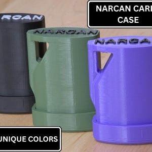 May include: Three Narcan carry cases in black, olive green, and purple. Each case features a top-mounted handle, a cylindrical body, and the word NARCAN embossed on the top surface. The text NARCAN CARRY CASE and 8 UNIQUE COLORS is visible.