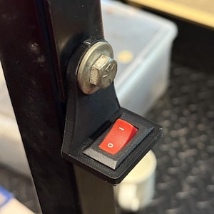May include: A black plastic switch holder with a red rocker switch, mounted on a black metal frame with a bolt and washer. The switch is labeled with "0" and "1" to indicate on and off positions.