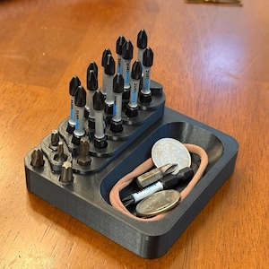 May include: A dark gray 3D-printed organizer with multiple compartments. It holds various screwdriver bits, coins, and a hair tie. The screwdriver bits are black and silver, and some have the brand name "toolant" printed on them.