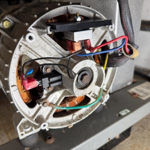 May include: Close-up of an electric motor with copper wiring, red, blue, and green wires, and a black component. The motor is housed in a silver metal casing with visible bolts and a black connector.