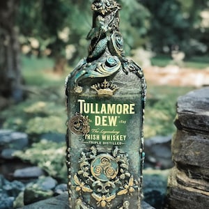 Puede incluir: Una botella ornamentada de whisky irlandés Tullamore D.E.W., triple destilado. La botella está decorada con adornos en color turquesa y dorado, incluyendo una sirena, diseños florales y el logotipo de la marca. La botella se exhibe al aire libre sobre una superficie de piedra.