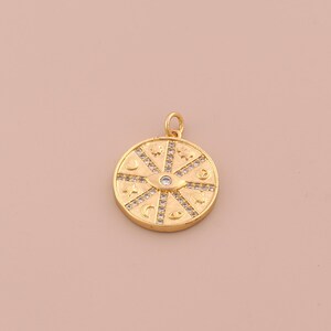 May include: Gold-colored pendant with a circular design. The pendant features an eye in the center, surrounded by various celestial symbols and small, clear stones. The pendant has a loop at the top for a chain.