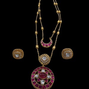 May include: A gold-toned jewelry set featuring a necklace with a large pendant and a pair of matching earrings. The pendant and earrings are adorned with red and clear gemstones. The necklace has a double strand design.