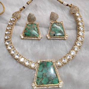 May include: A gold necklace and earring set. The necklace and earrings feature large, green, rectangular stones with a marbled appearance, surrounded by small, clear stones. The necklace has a series of clear, oval-shaped stones along the band.