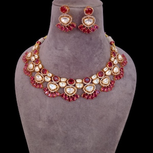 Puede incluir: Un conjunto de collar y pendientes dorados. El collar y los pendientes presentan una combinación de piedras preciosas rojas y claras. El collar tiene un diseño festoneado con piedras rojas y claras alternas. Los pendientes tienen un diseño similar.