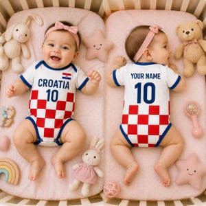 May include: Two babies wearing white onesies with red and white chequered patterns and blue trim. One onesie says "CROATIA 10" and the other says "YOUR NAME 10". They are lying on pink bedding with stuffed animals and toys.