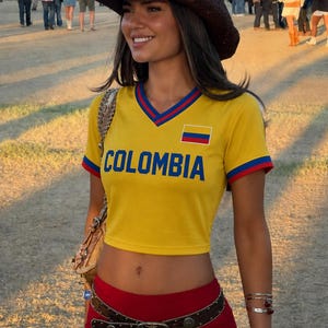 May include: A woman wearing a yellow crop top with "COLOMBIA" printed in blue, a red skirt, and a brown cowboy hat. The top has a V-neck with blue and red trim and a small Colombian flag. A brown belt with silver studs and a brown purse complete the look.