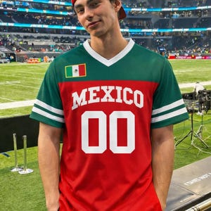 Może przedstawiać: Czerwona, zielona i biała koszulka Meksyku z napisem "MEXICO" białymi literami blokowymi i numerem "00". Koszulka ma dekolt w serek i białe paski na rękawach. Osoba ma na sobie czerwoną czapkę z daszkiem.