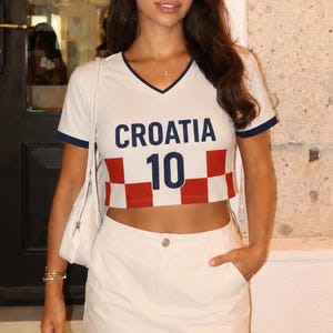 May include: White crop top with navy blue trim and the word "CROATIA" in navy blue letters. Below the word "CROATIA" is the number "10" in navy blue. The bottom of the shirt has a red and white checkered pattern. The shirt is paired with a white skirt.