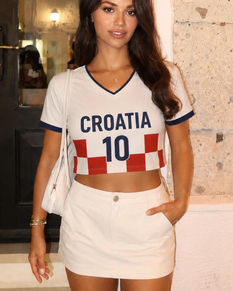 May include: White crop top with navy blue trim and the word "CROATIA" in navy blue letters. Below the word "CROATIA" is the number "10" in navy blue. The bottom of the shirt has a red and white checkered pattern. The shirt is paired with a white skirt.