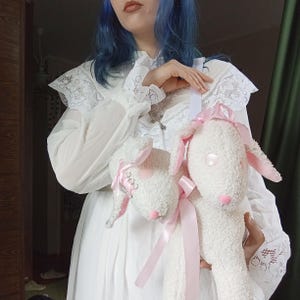 以下が含まれることがあります： ピンクのアクセントとリボンが付いた、2つのぬいぐるみの子羊のおもちゃ。子羊は白くふわふわの体、ピンクの耳、ピンクの鼻をしています。1匹の子羊の耳には銀色の装飾が施されています。おもちゃは白いドレスの前に置かれています。